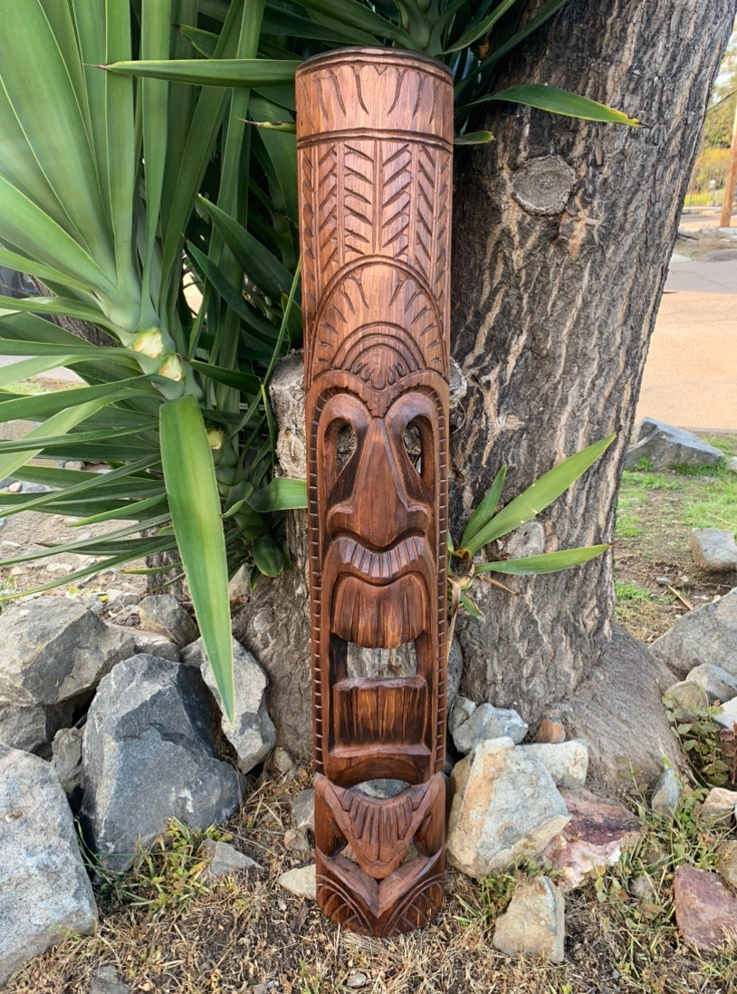 Carved wooden tiki totem pole leaning against a tree outdoors in a garden.
