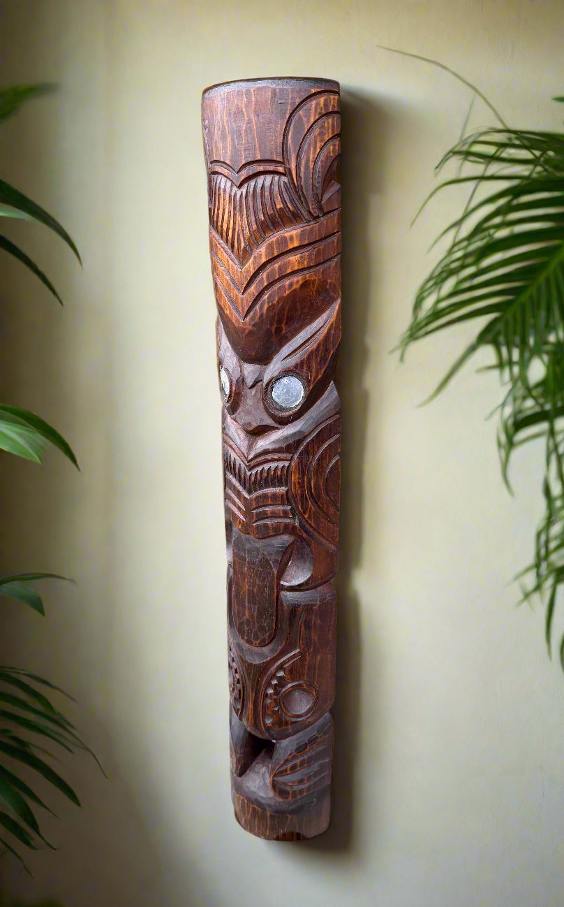 Tall carved wooden tiki totem pole with geometric patterns and a stylized face against a pale wall.