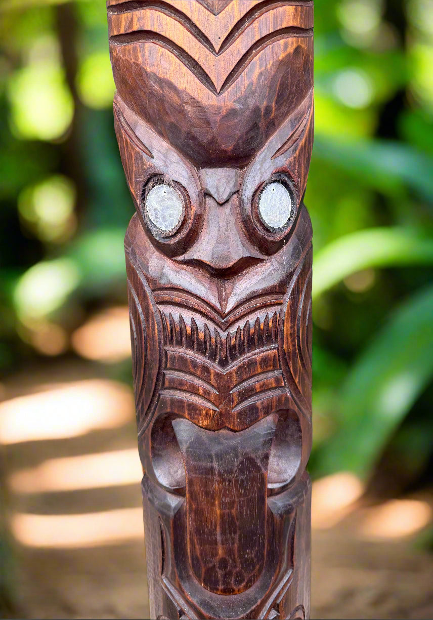 Carved wooden tiki totem with round eyes and intricate patterns in a tropical garden.