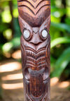 Carved wooden tiki totem with round eyes and intricate patterns in a tropical garden.