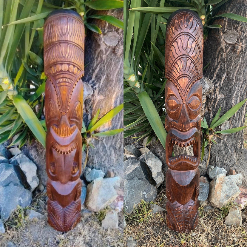 Two-sided view of a tall carved wooden tiki totem statue outdoors among rocks and plants.