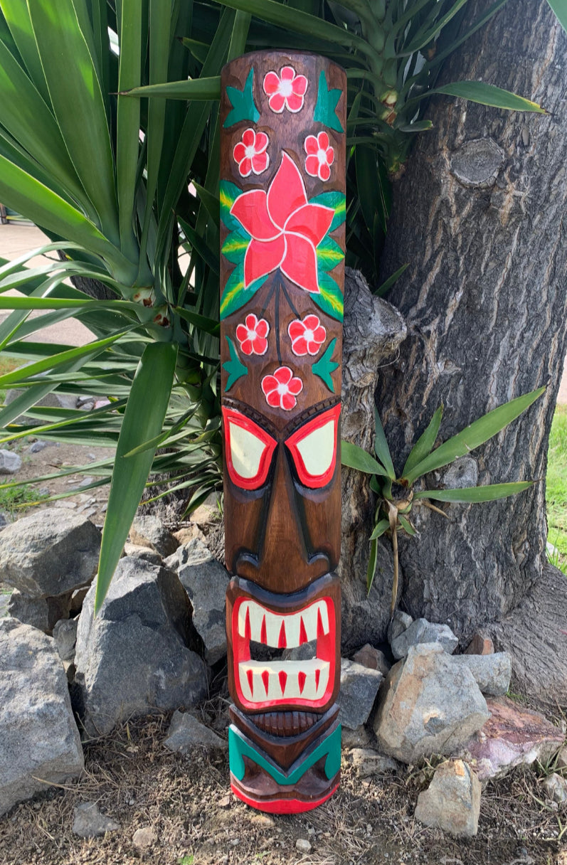 Carved wooden tiki statue with red eyes and teeth, hibiscus flower patterns, set among rocks and tropical plants.