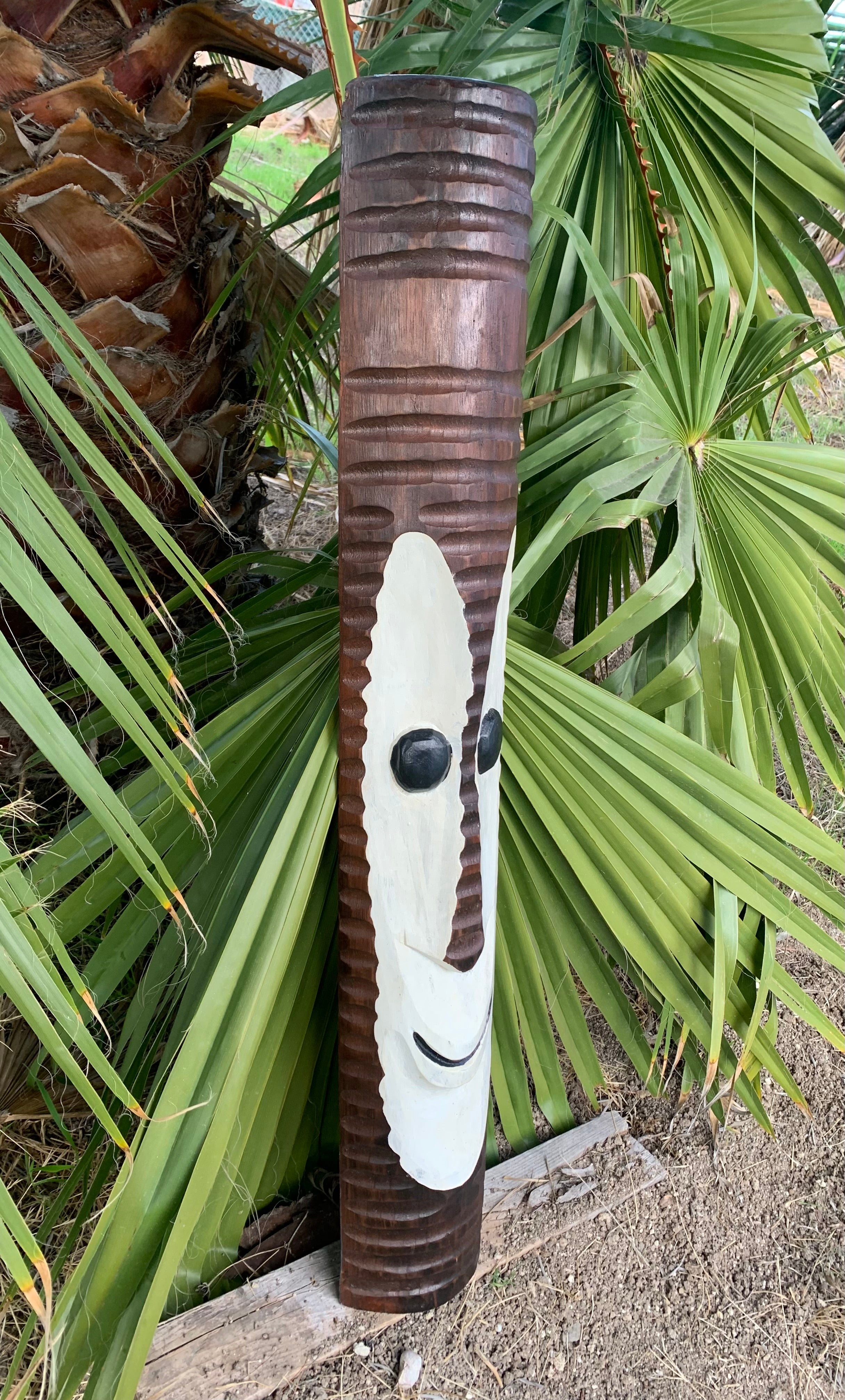 Carved wooden totem pole with a white face and black eyes beside palm leaves