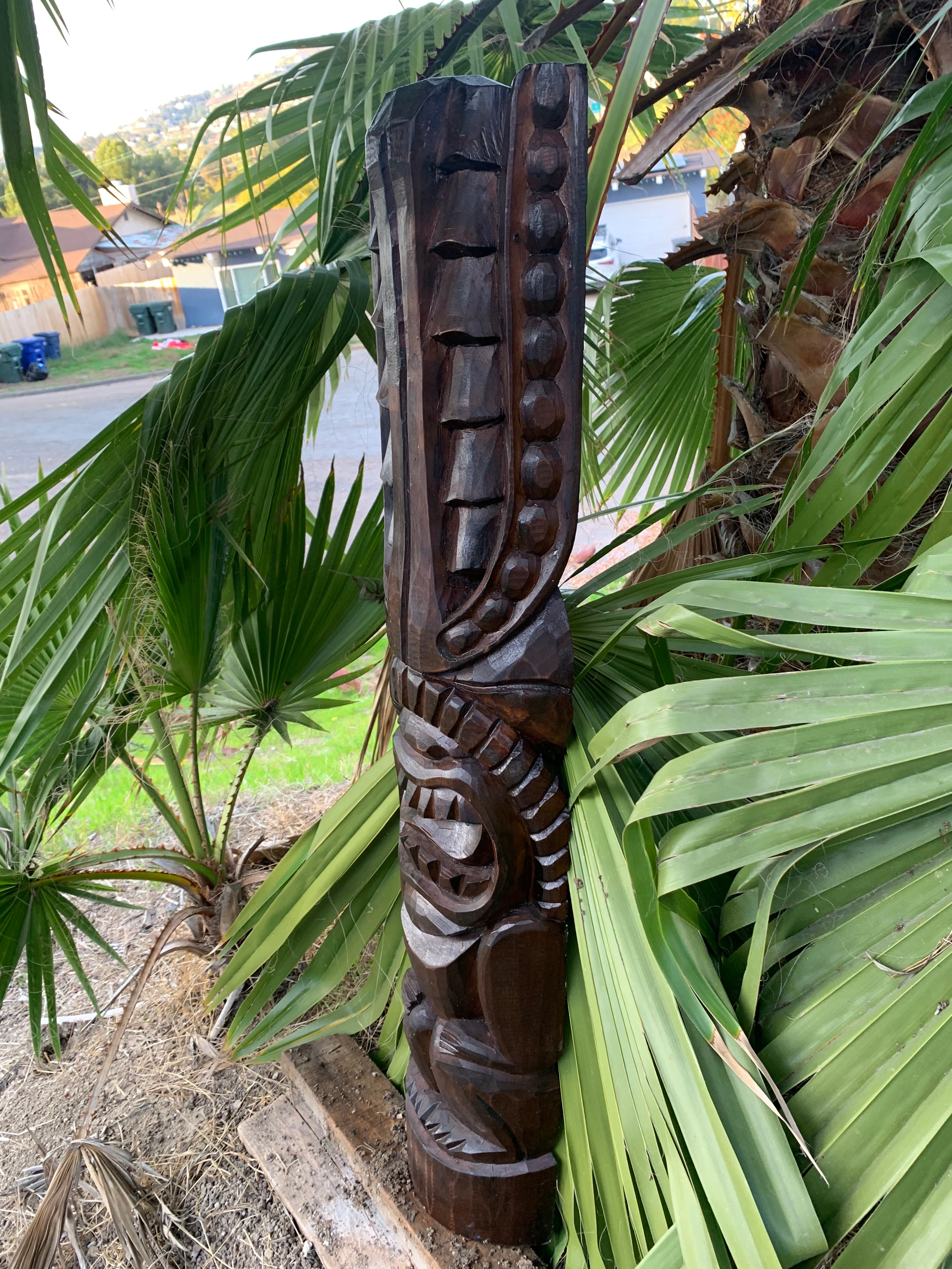 Carved dark wood totem pole with circular bead motifs among palm fronds in a yard.
