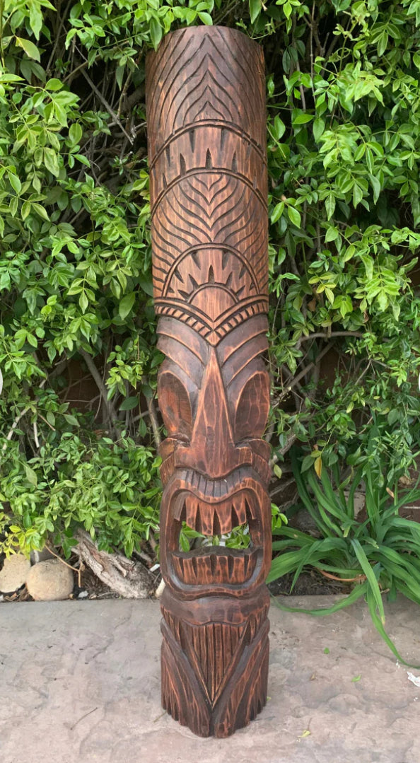Carved wooden totem pole with geometric patterns outdoors among green bushes.