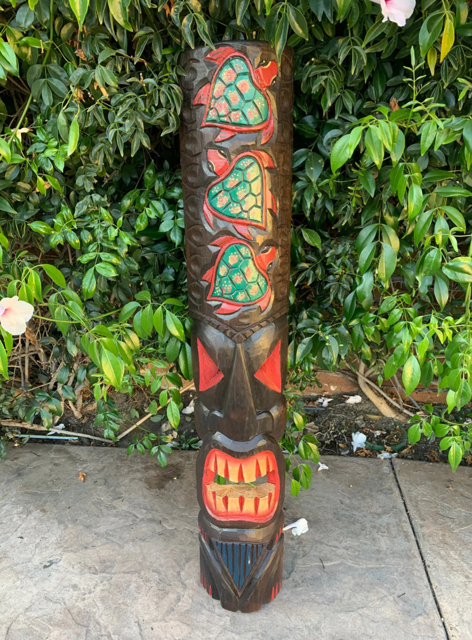 Carved wooden totem pole with three turquoise turtle motifs among lush garden greenery