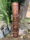 Carved wooden totem pole featuring a tiki face, outdoors beside a tree and rocks