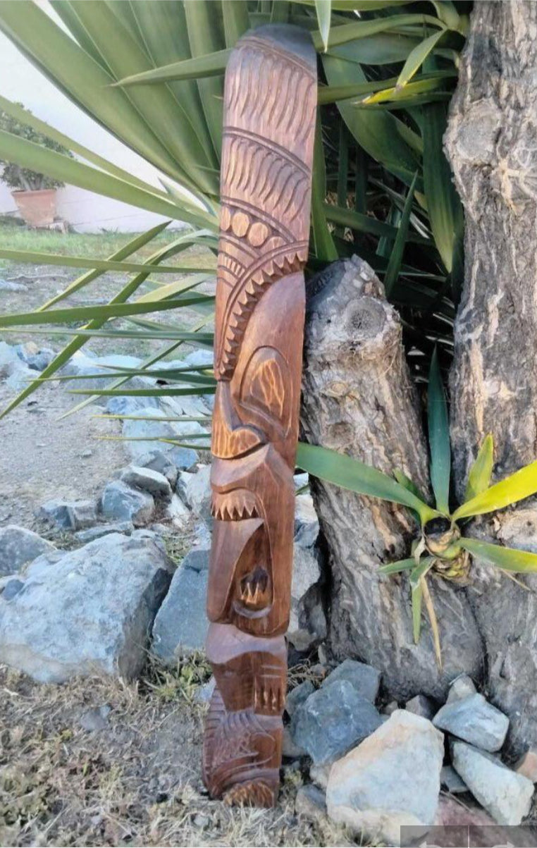 Tall wooden totem pole with tribal geometric patterns and carved faces outdoors among rocks and plants