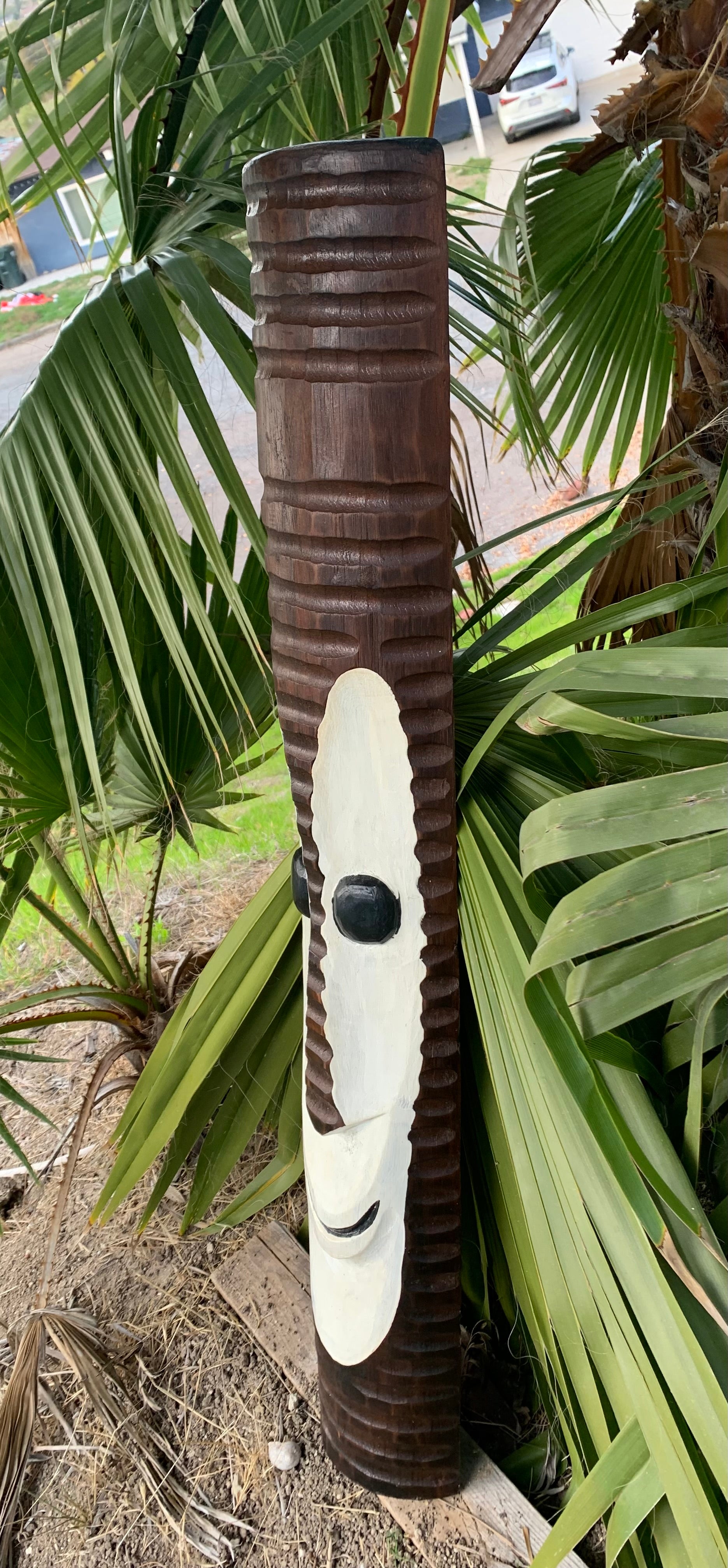 Brown wooden totem pole with a white face carving among palm leaves outdoors