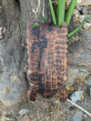 Charred wood plaque with scalloped edges hanging on a tree trunk with a green plant in the background.
