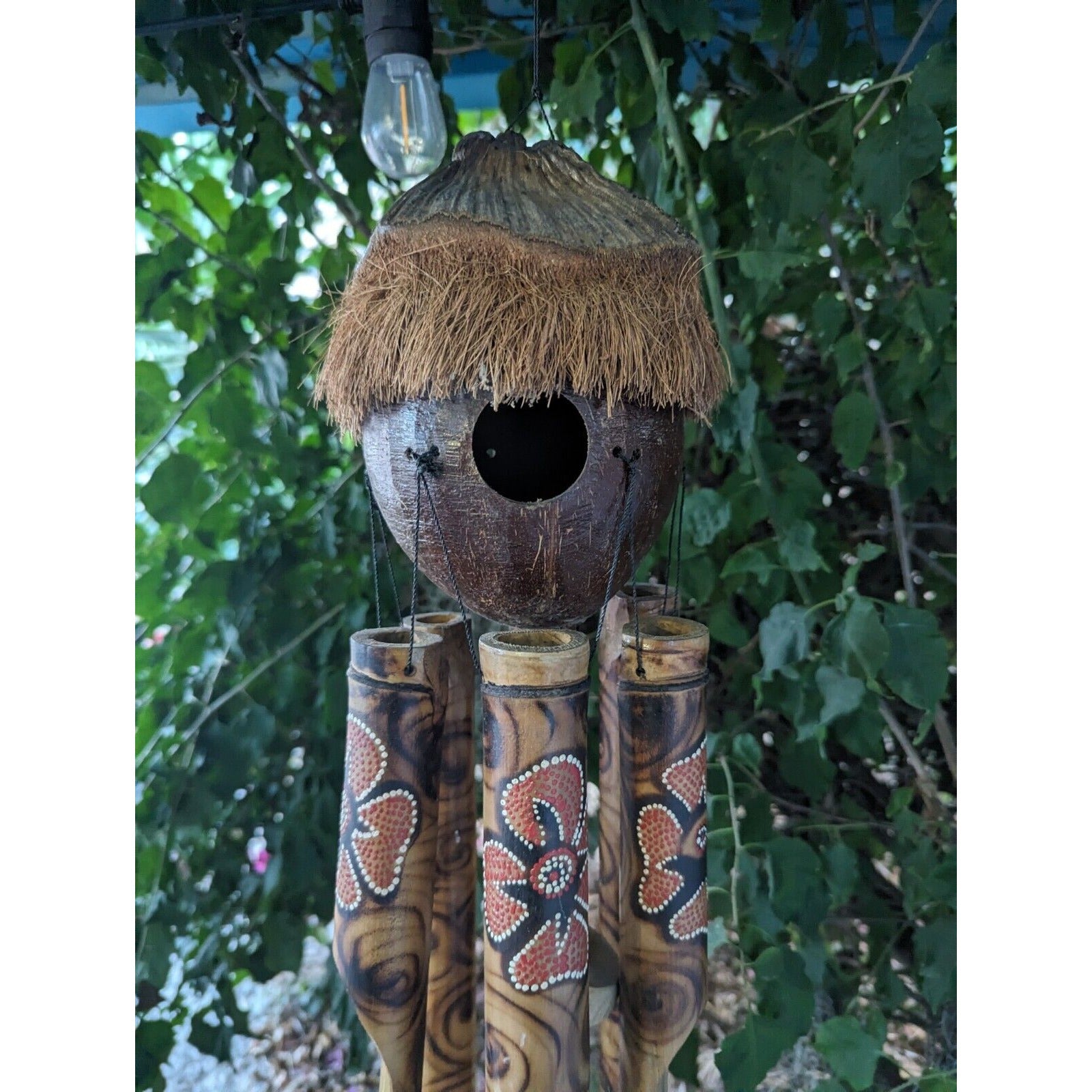 Coconut-shell birdhouse with straw thatch roof and hanging bamboo chimes in a garden setting