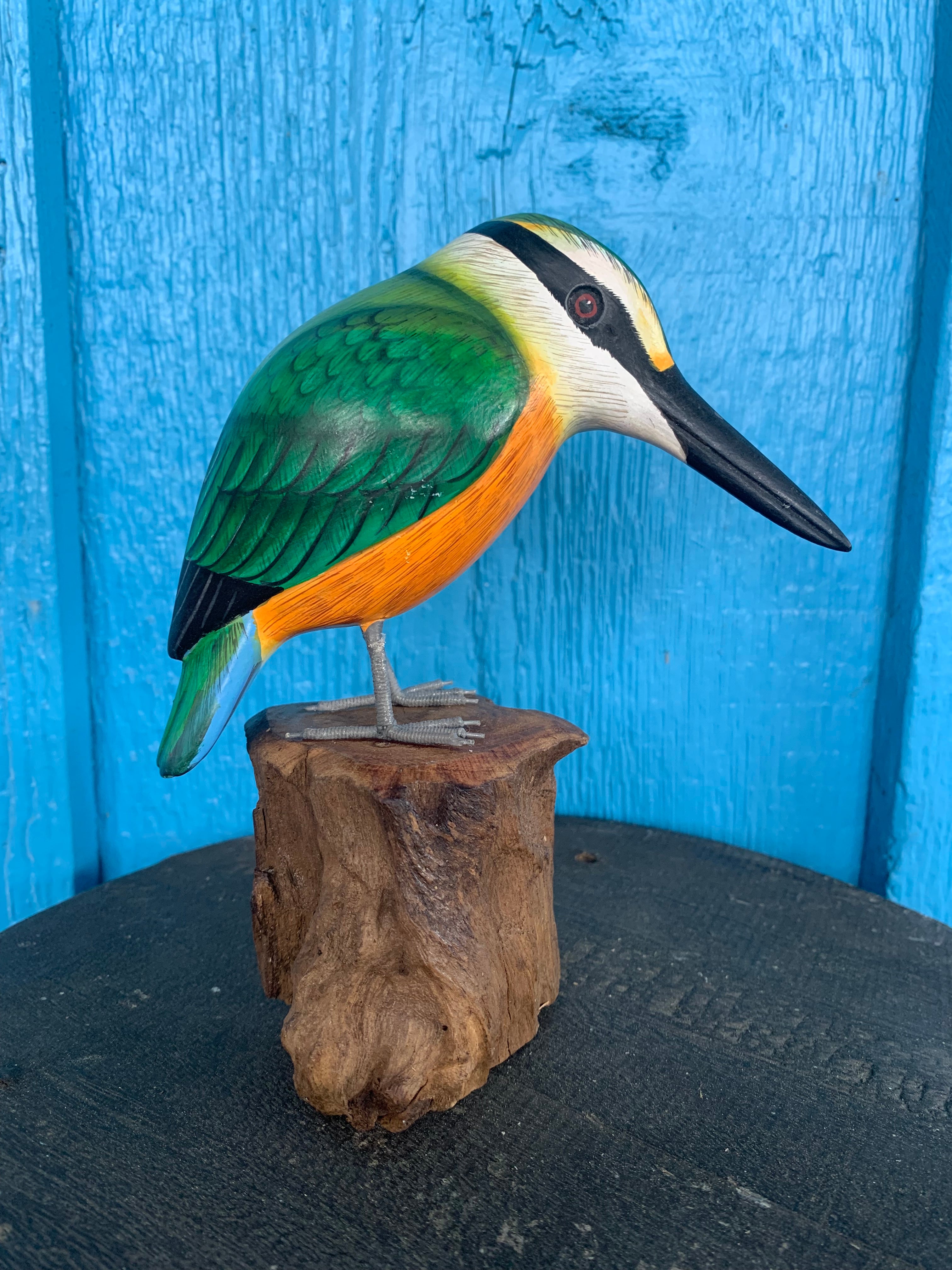 Colorful carved wooden bird statue with green wings and orange belly perched on a wooden stump against a blue background