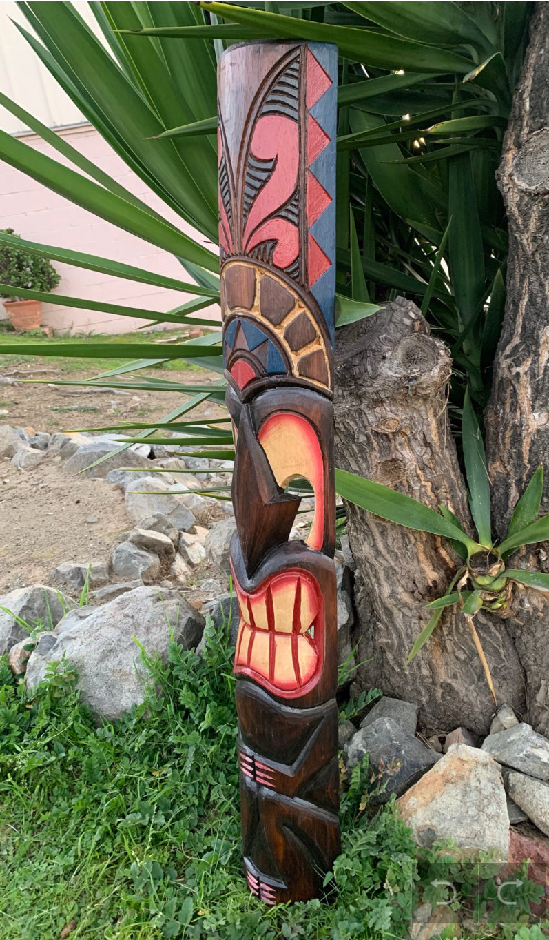 Tall, colorfully carved wooden totem pole with geometric patterns beside greenery and rocks