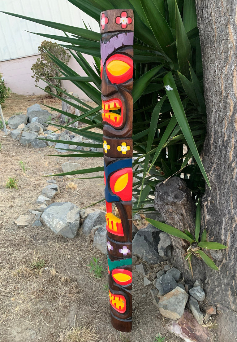 Colorful carved totem pole with flower motifs standing outdoors beside a tree and rocks