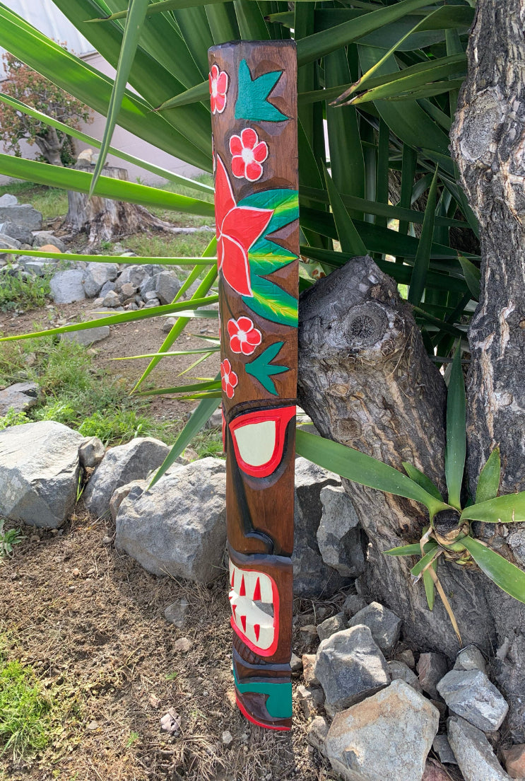 Colorful carved wooden totem pole with red flowers and teal leaves in a garden setting