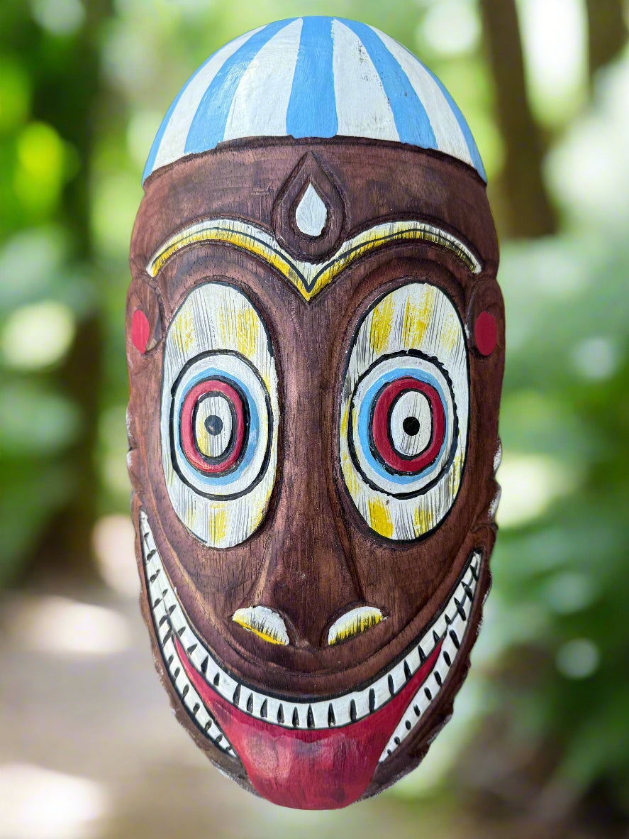Colorful carved wooden mask with large round eyes and a wide toothy grin in a forest setting
