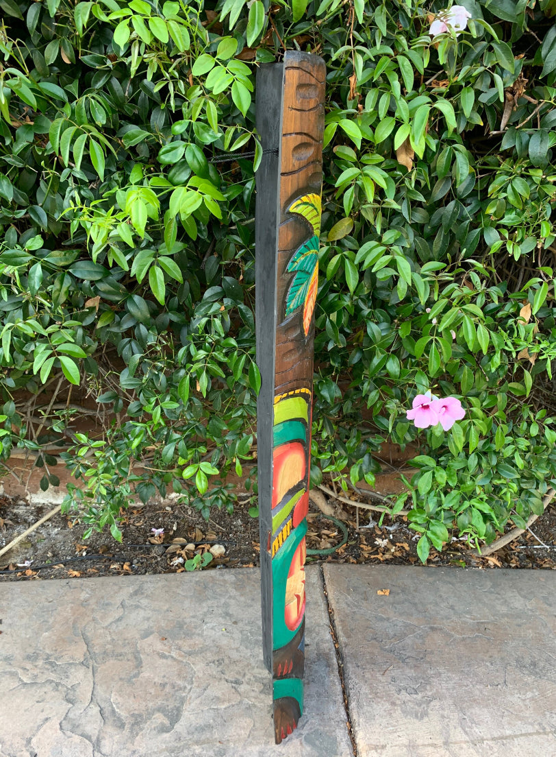 Colorful carved wooden totem pole standing beside a dense green hedge with pink flowers nearby.