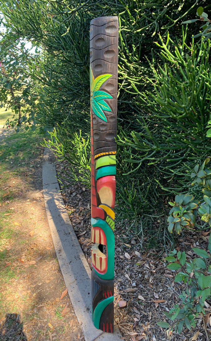 Colorful carved wooden totem pole with leaf design beside a hedge along a path