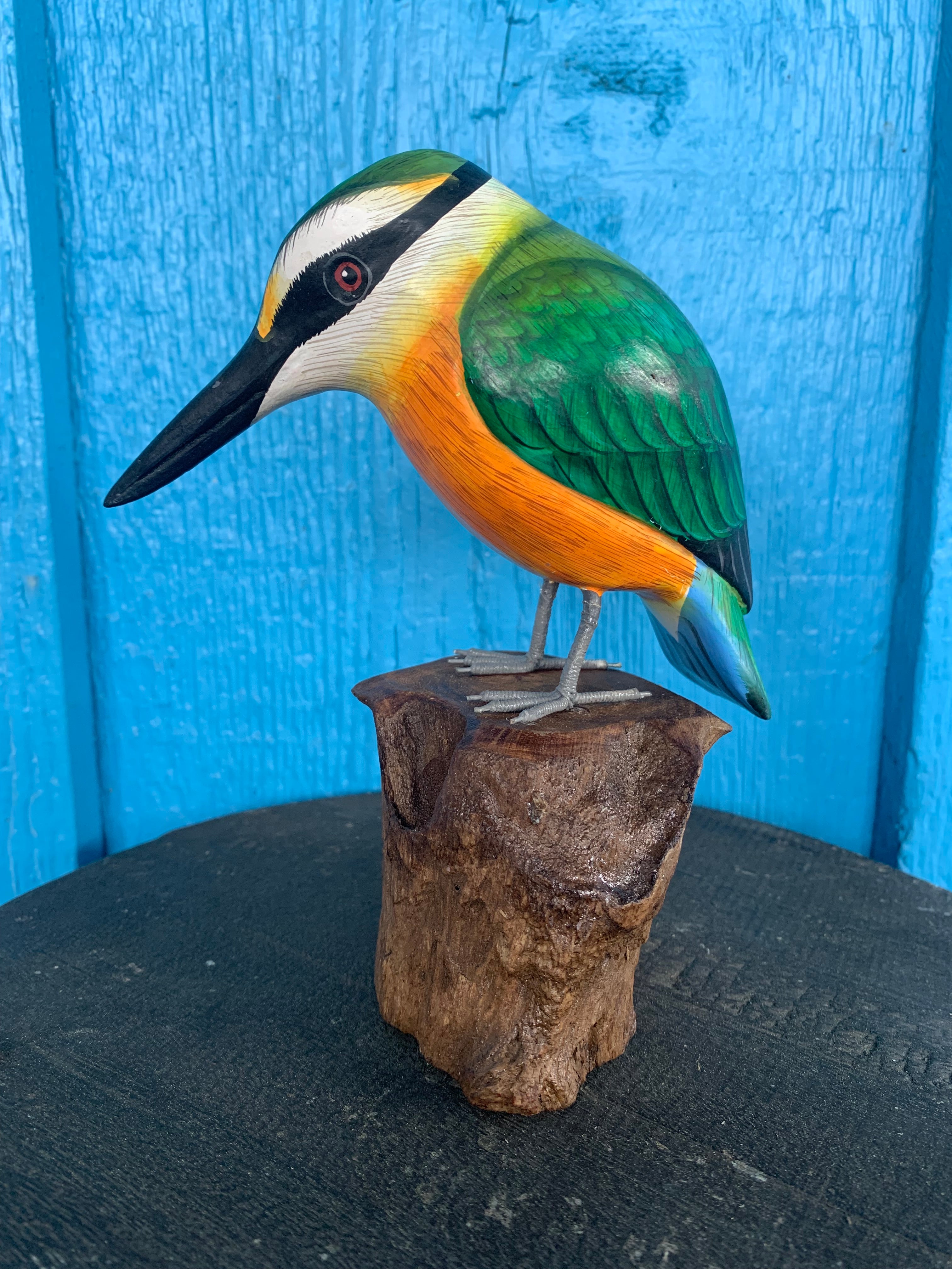Colorful kingfisher sculpture perched on a wooden stump against a blue background.