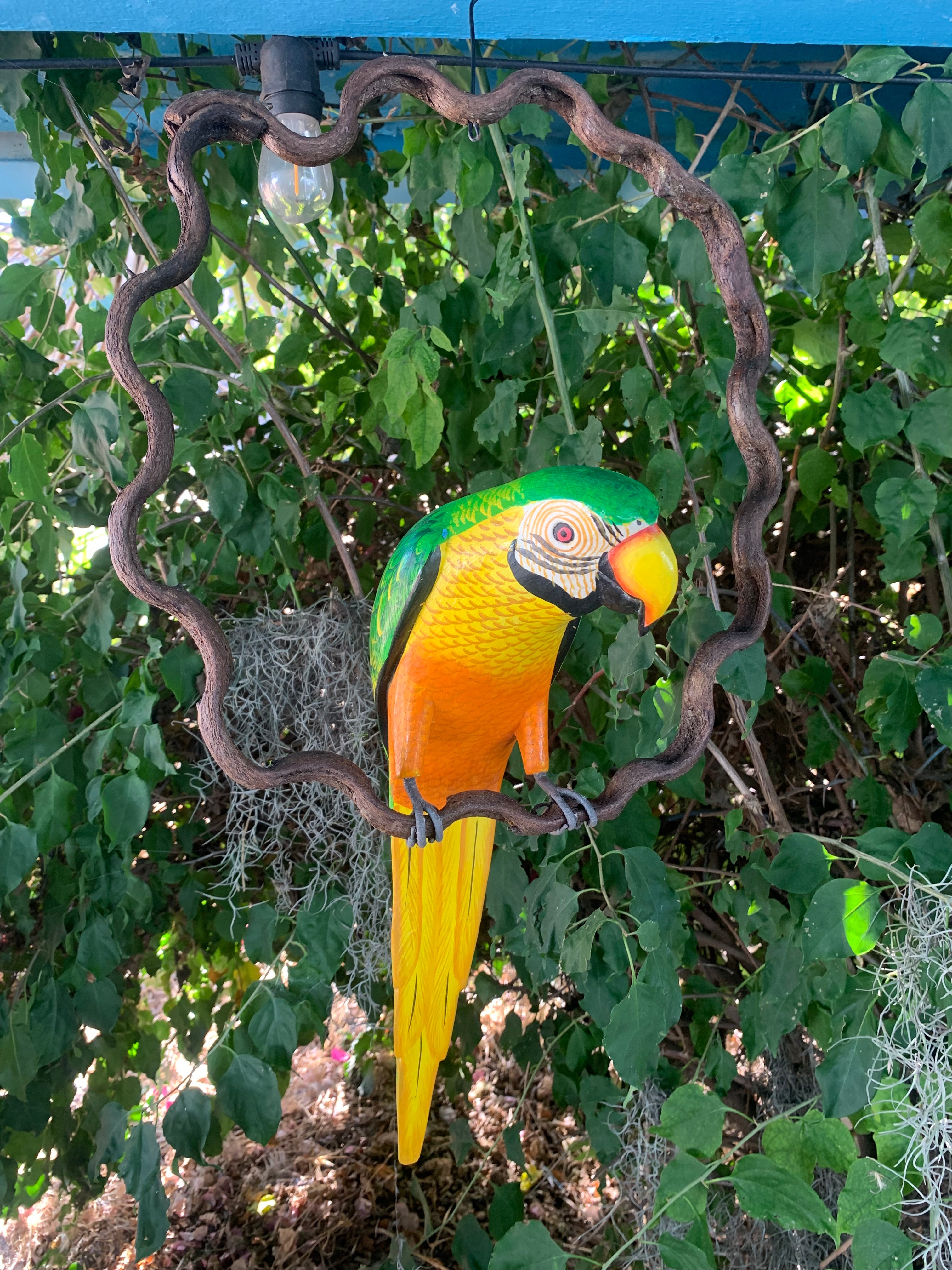 Colorful macaw parrot sculpture in green and yellow perched on a wavy brown ring with leafy background.