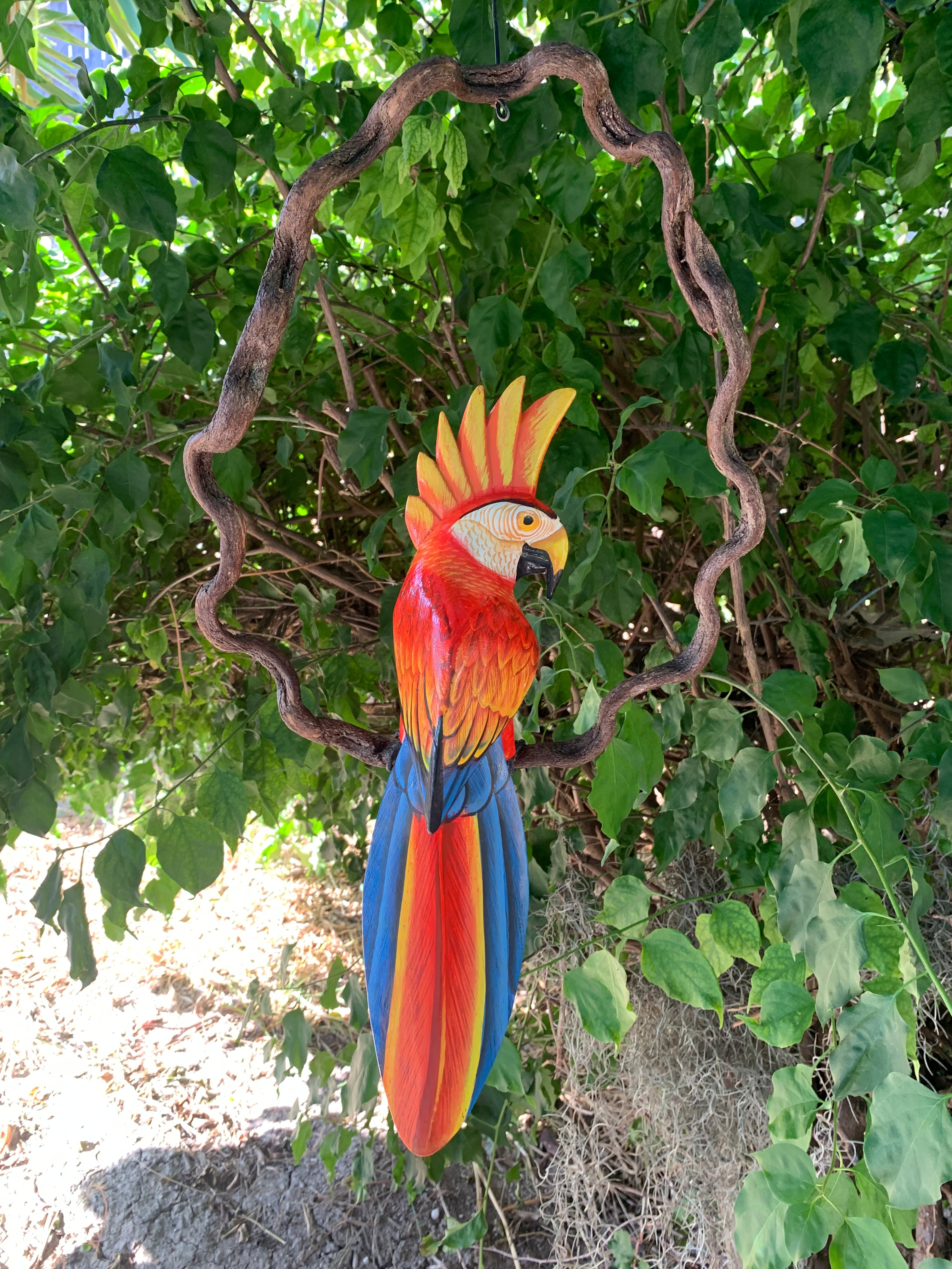 Colorful macaw parrot sculpture hanging in a leafy garden with a twisted circular branch frame.