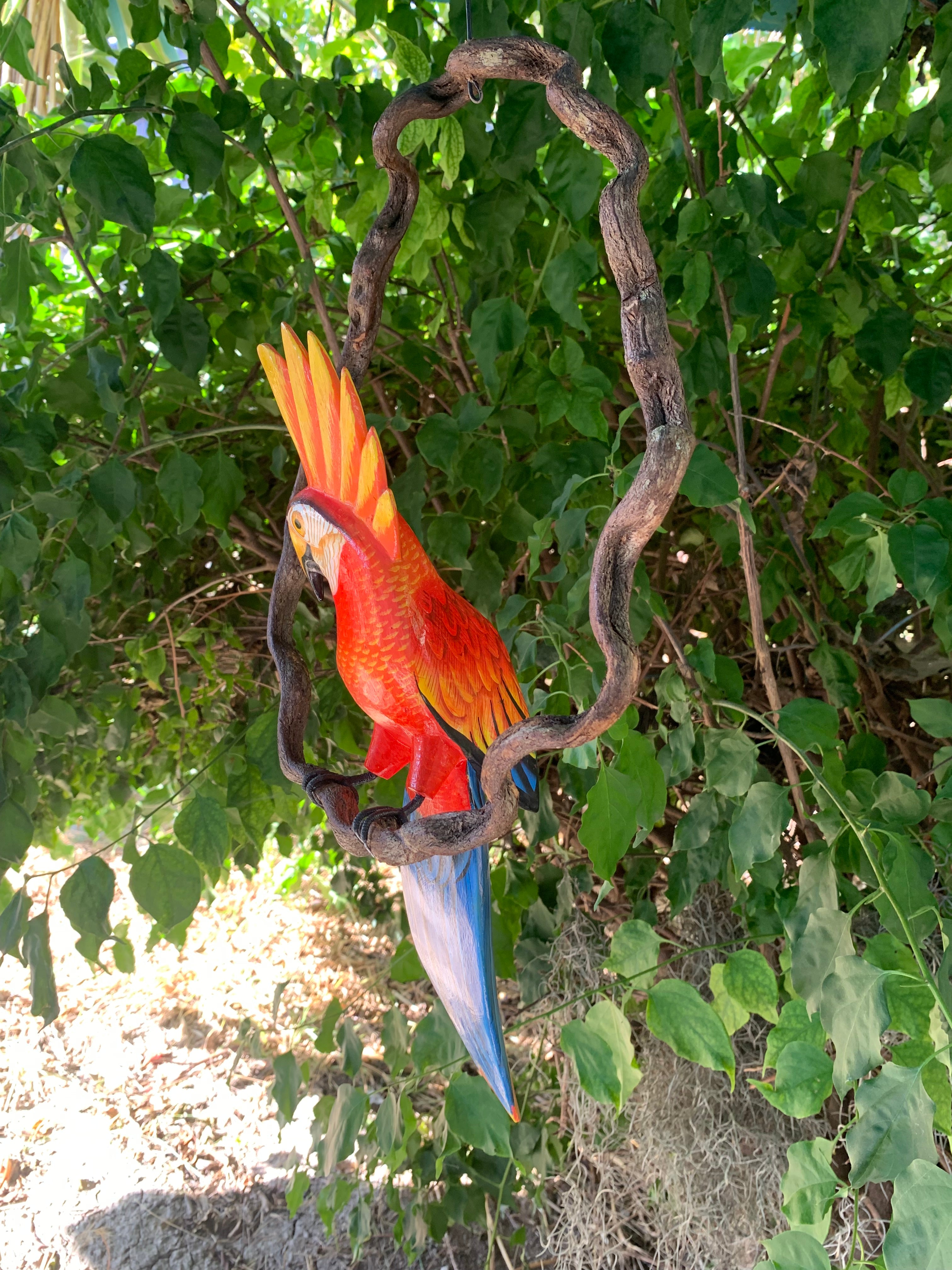 Colorful macaw sculpture with orange crest and blue tail hanging from a twisted branch among green leaves.