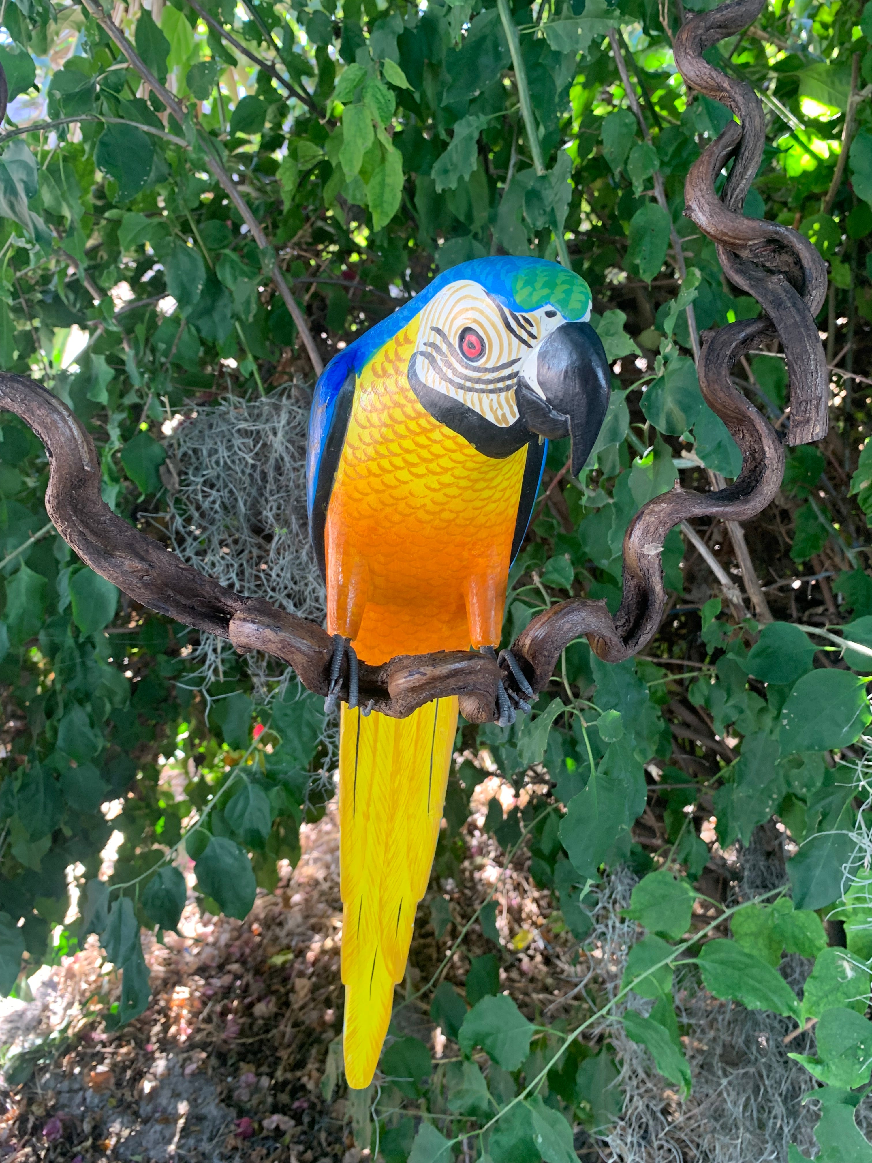 Colorful macaw sculpture with blue, yellow, and orange feathers perched on a twisted branch among green foliage.