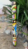 Tall painted totem pole with bright geometric shapes and white flowers, set outdoors among rocks and plants.