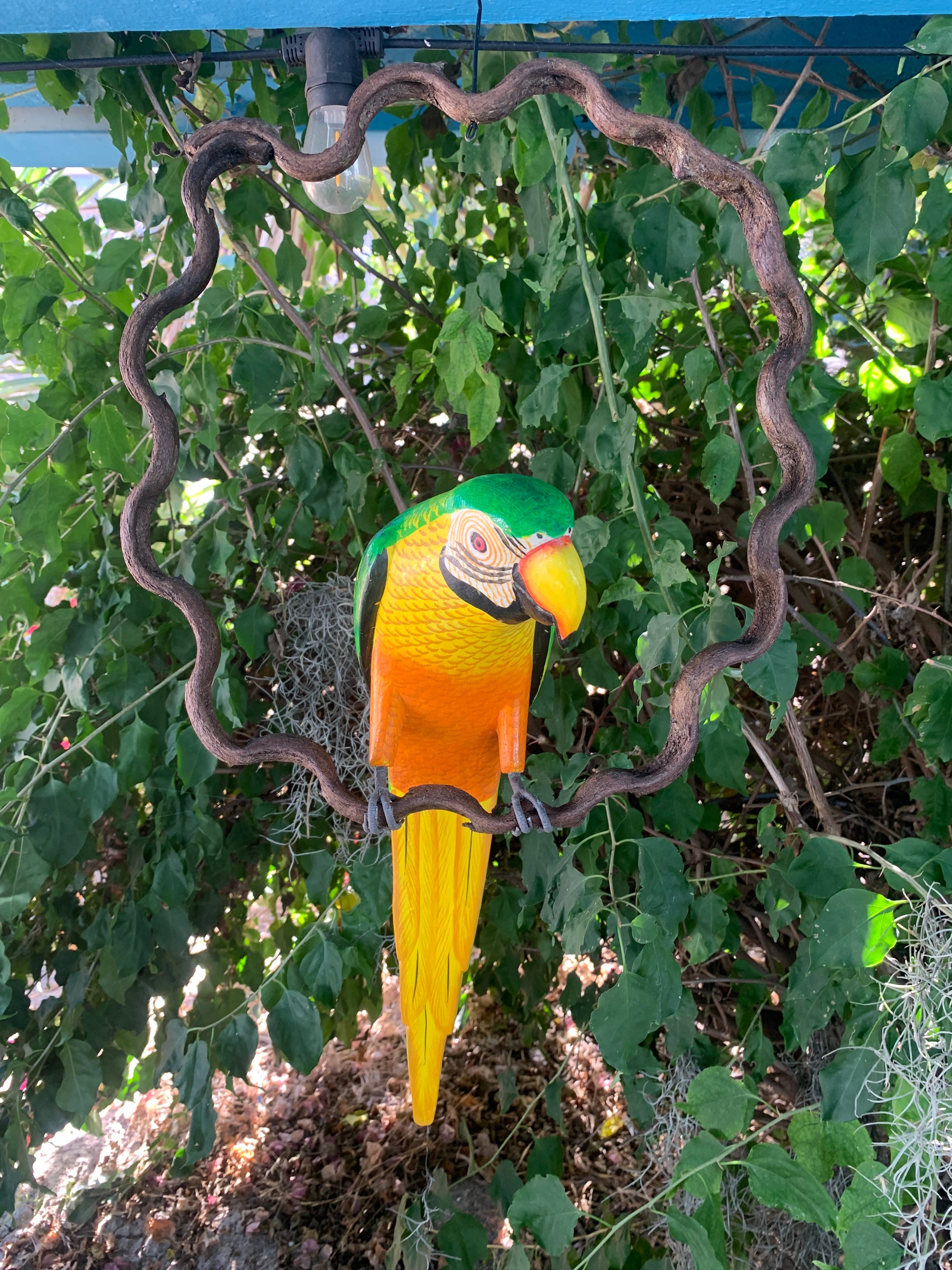 Colorful parrot garden sculpture with green head, orange body and long yellow tail in a leafy setting