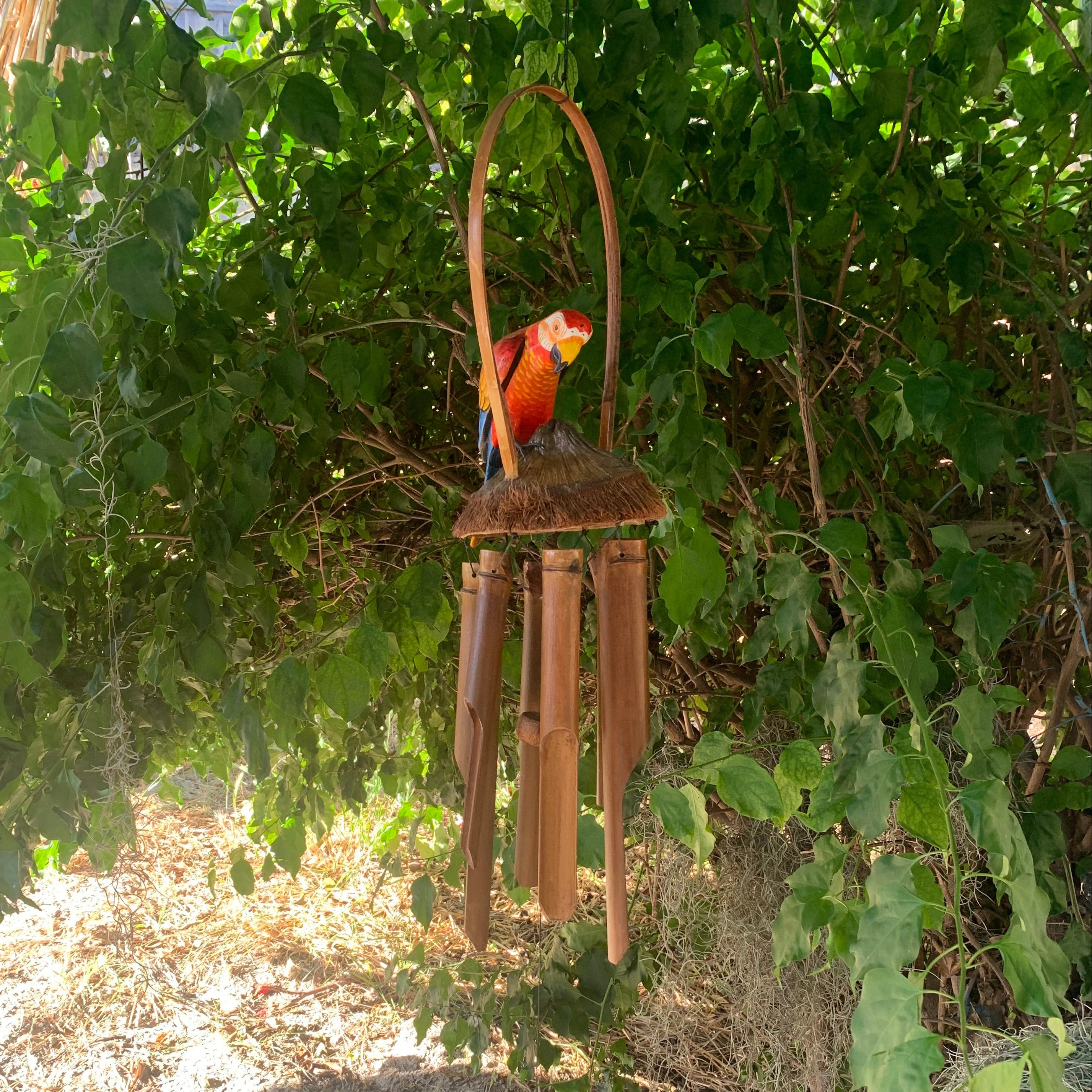 Colorful parrot wind chime hanging from a vine-covered bush in a garden
