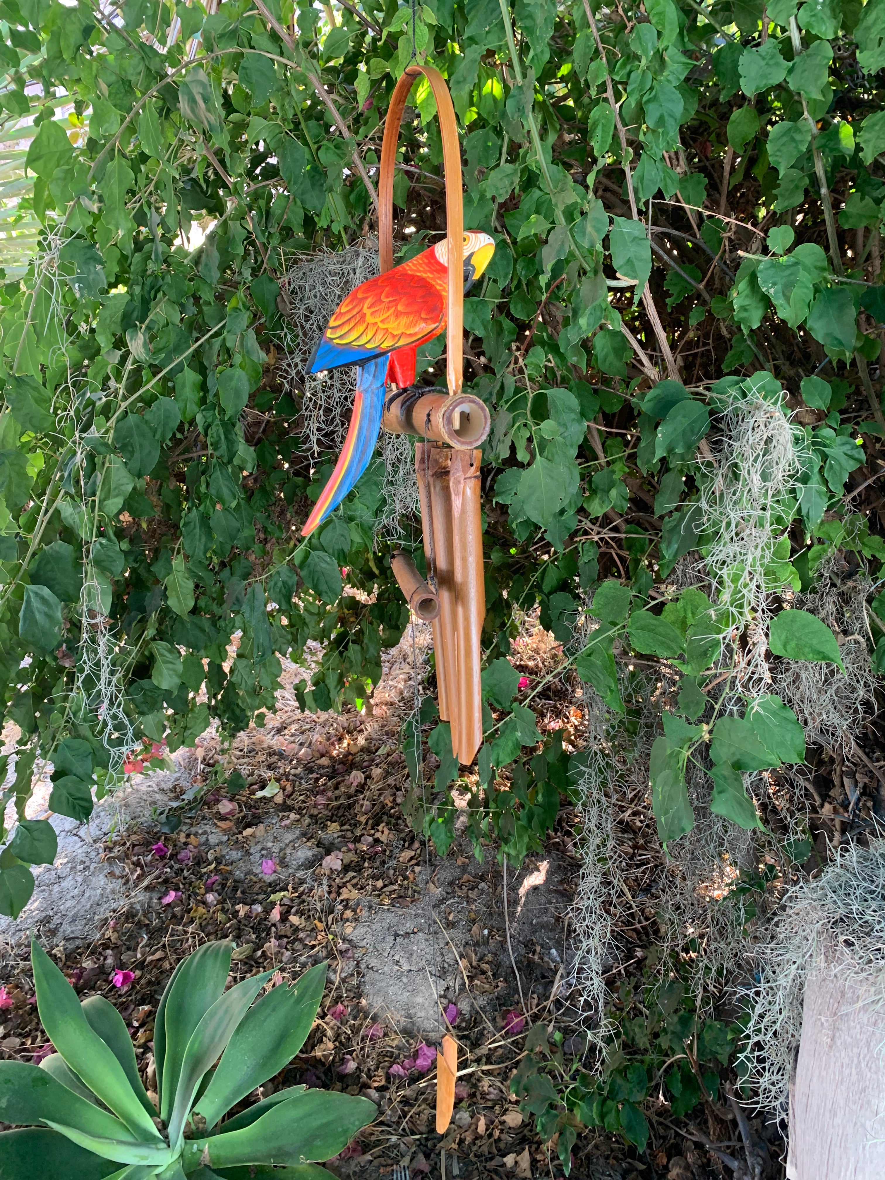 Colorful parrot wind chime perched on a bamboo stand in a leafy garden.