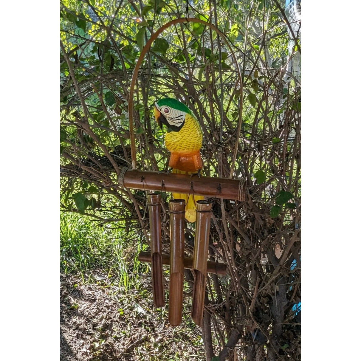 Colorful wooden parrot statue perched on bamboo wind chimes in a garden.