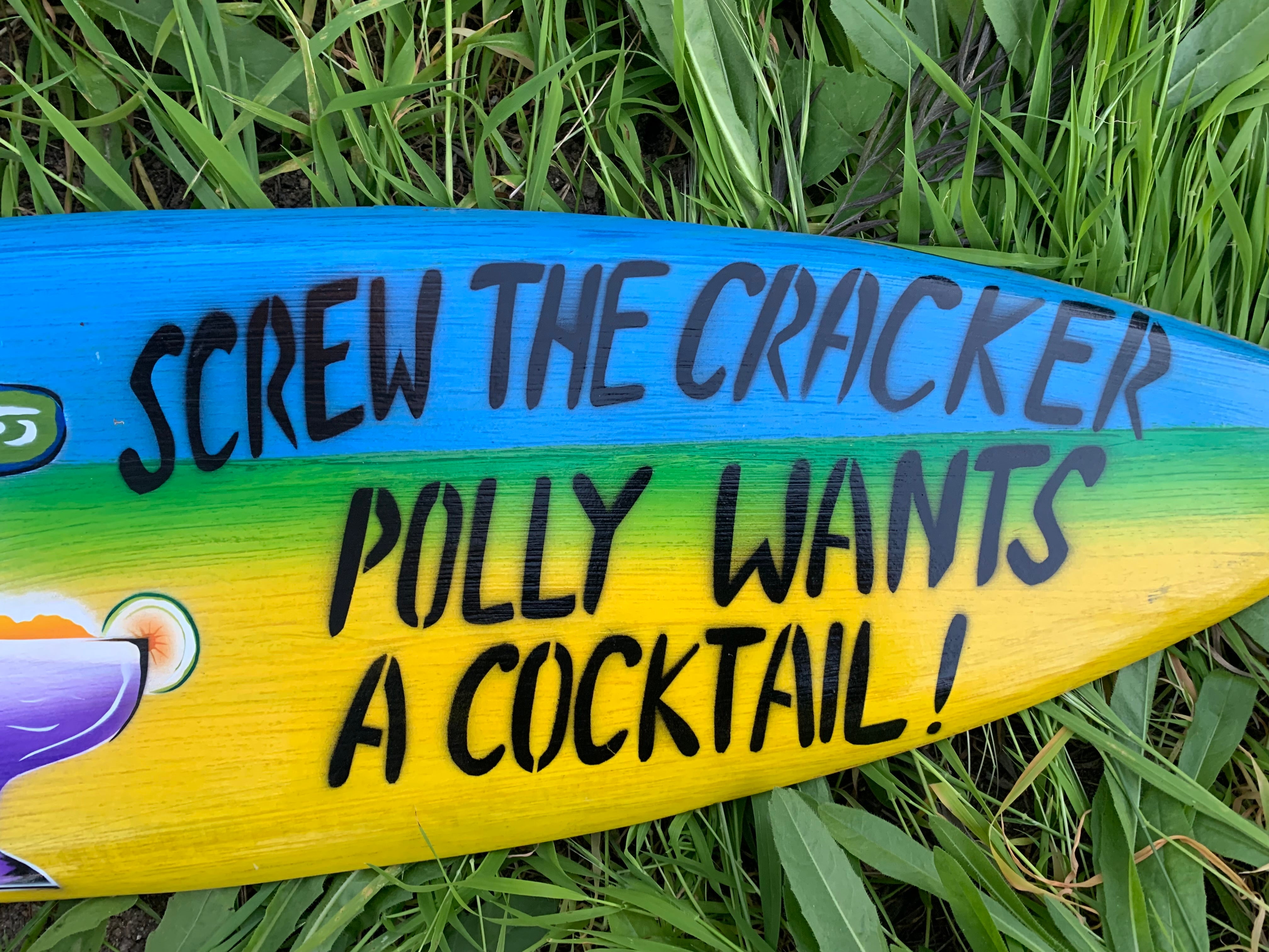 Rainbow-colored surfboard with bold black text reading 'Screw the cracker Polly wants a cocktail' on green grass