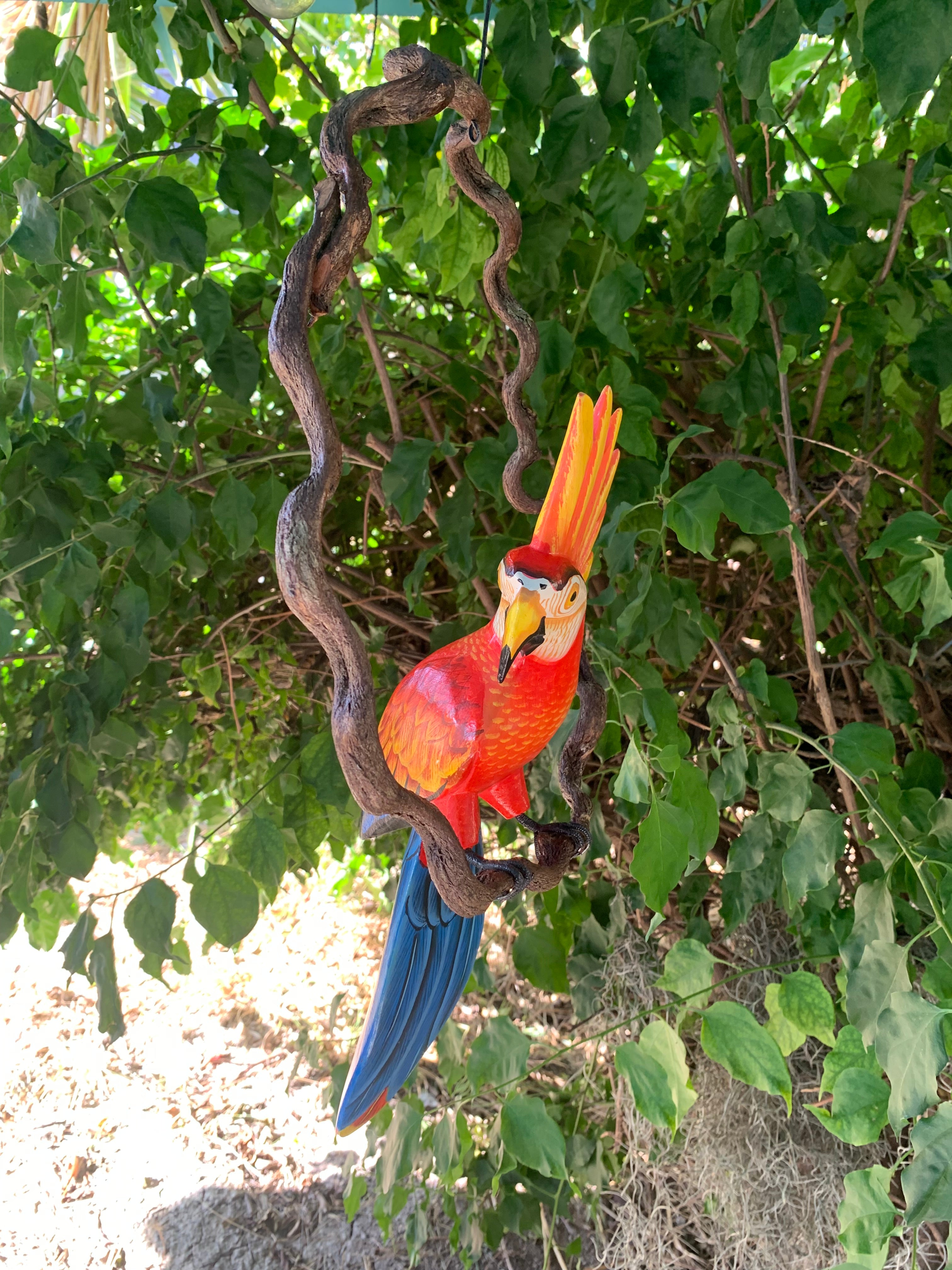 Colorful red macaw parrot sculpture hanging on twisting brown vine with green leaves in background