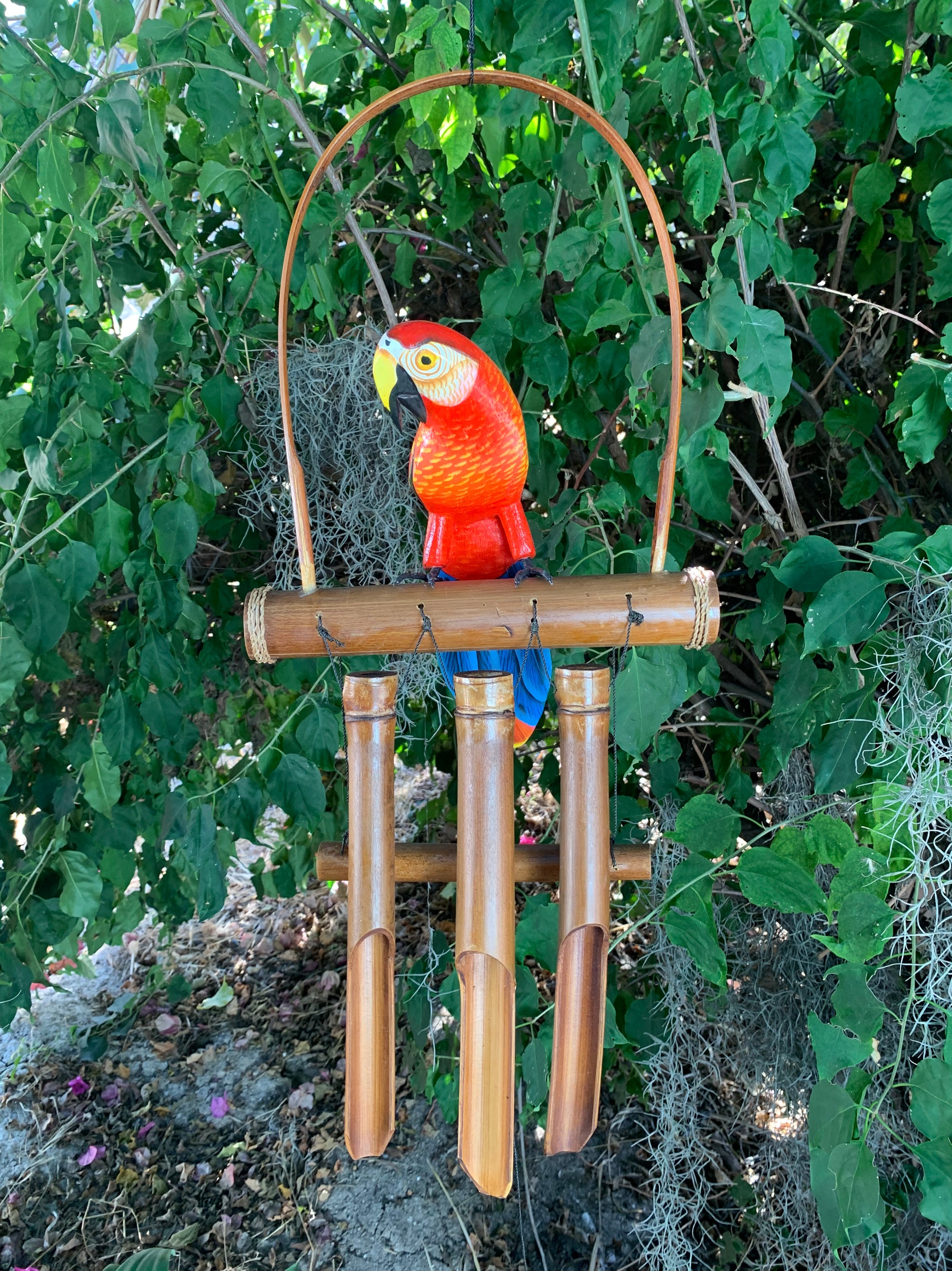 colorful red parrot sculpture perched on a wooden wind chime with bamboo tubes among green garden foliage