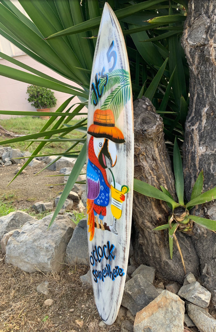 Colorful tropical surfboard featuring a bird wearing a hat among palm leaves with the phrase 5 oclock somewhere