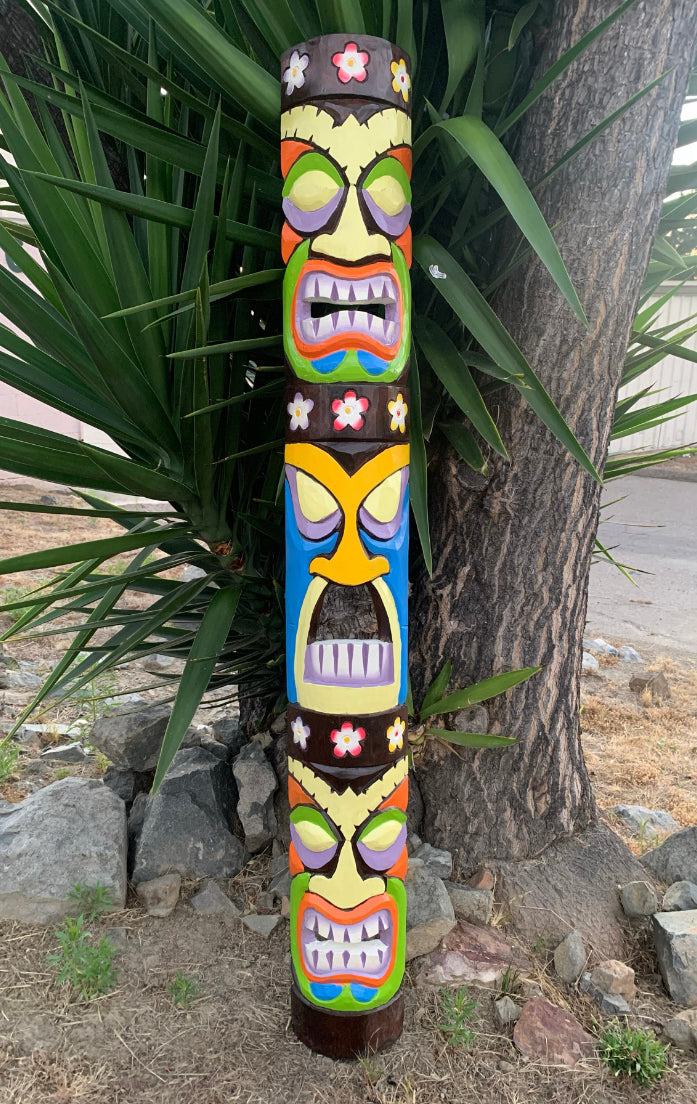 Colorful tiki totem pole with carved faces outdoors beside a tree