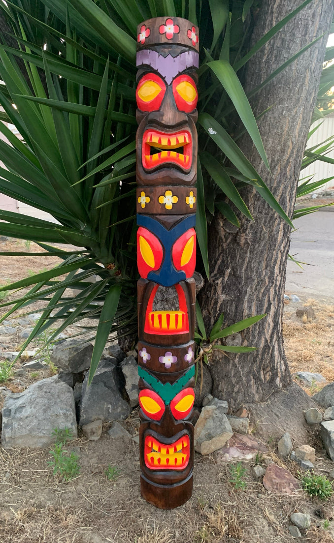 Colorful wooden tiki totem pole with multiple carved faces outdoors beside a tree