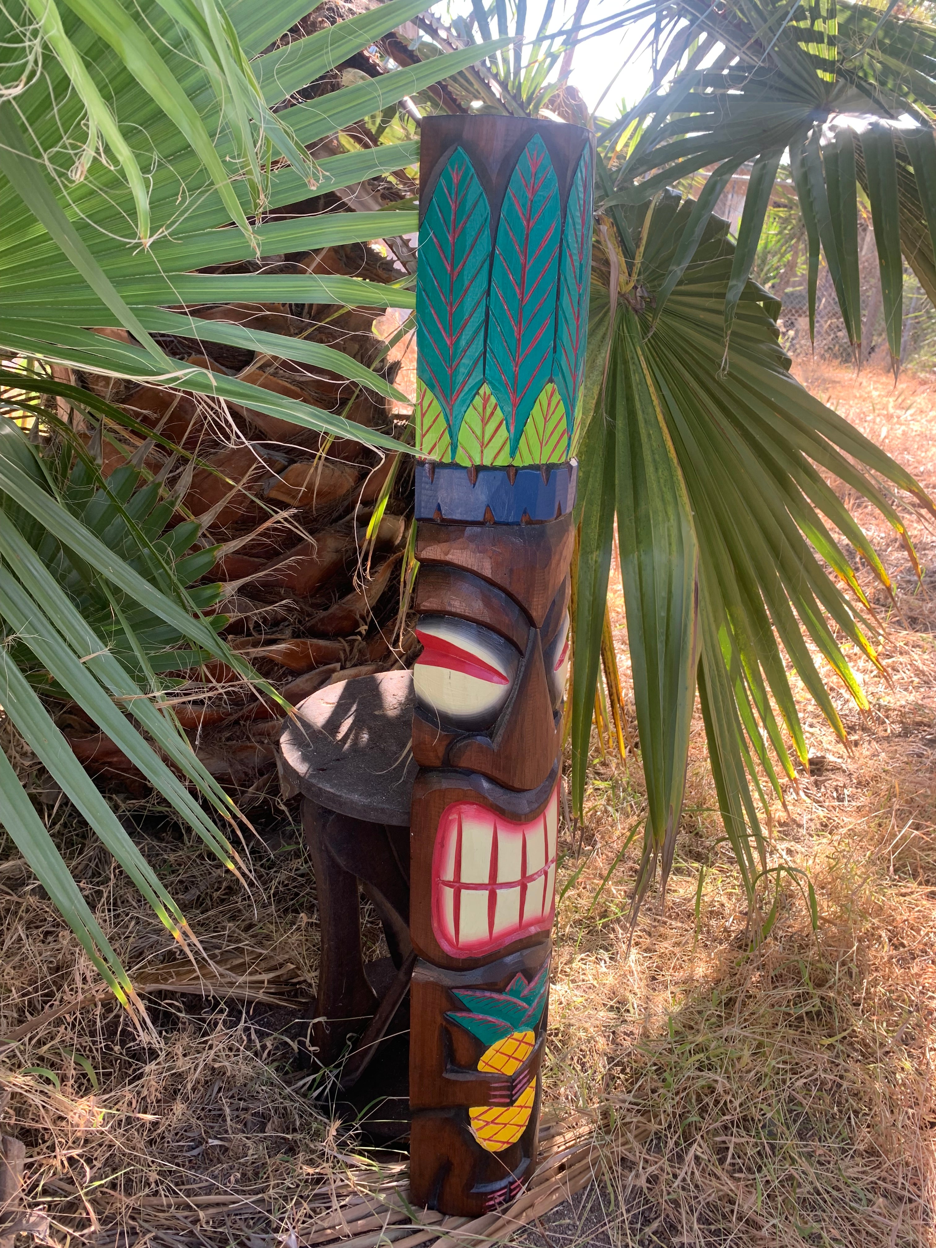 Colorful carved tiki totem pole nestled among palm fronds with leaf-patterned top and a wide grinning mouth.