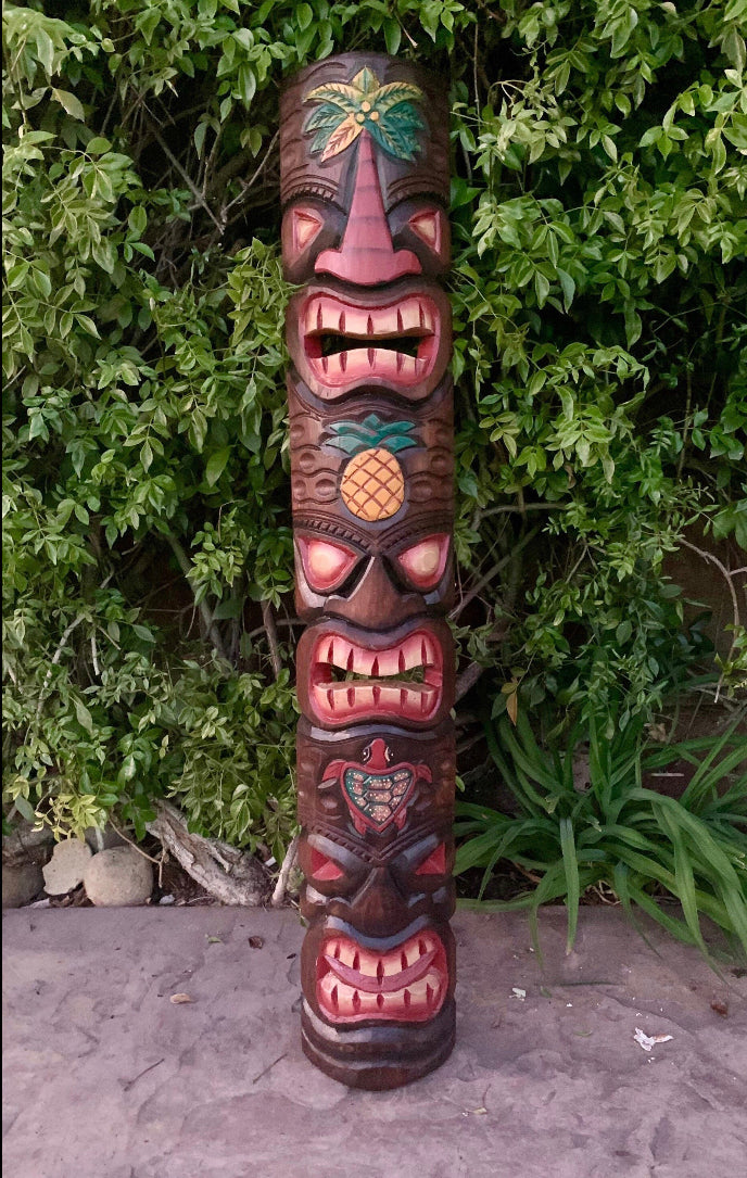 colorful wooden tiki-totem-pole with multiple carved faces and a pineapple motif in a leafy garden