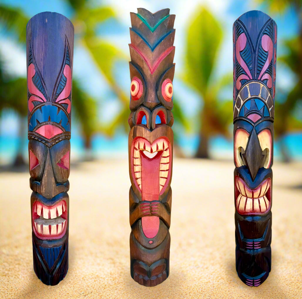 Three colorful carved tiki poles stand upright on a sandy beach with blurred palm trees behind.