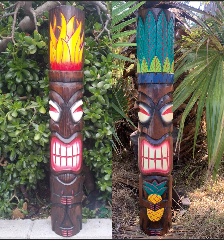 Two colorful carved tiki statues with expressive faces; left features a flame crest, right has a feather motif and pineapple at the base.