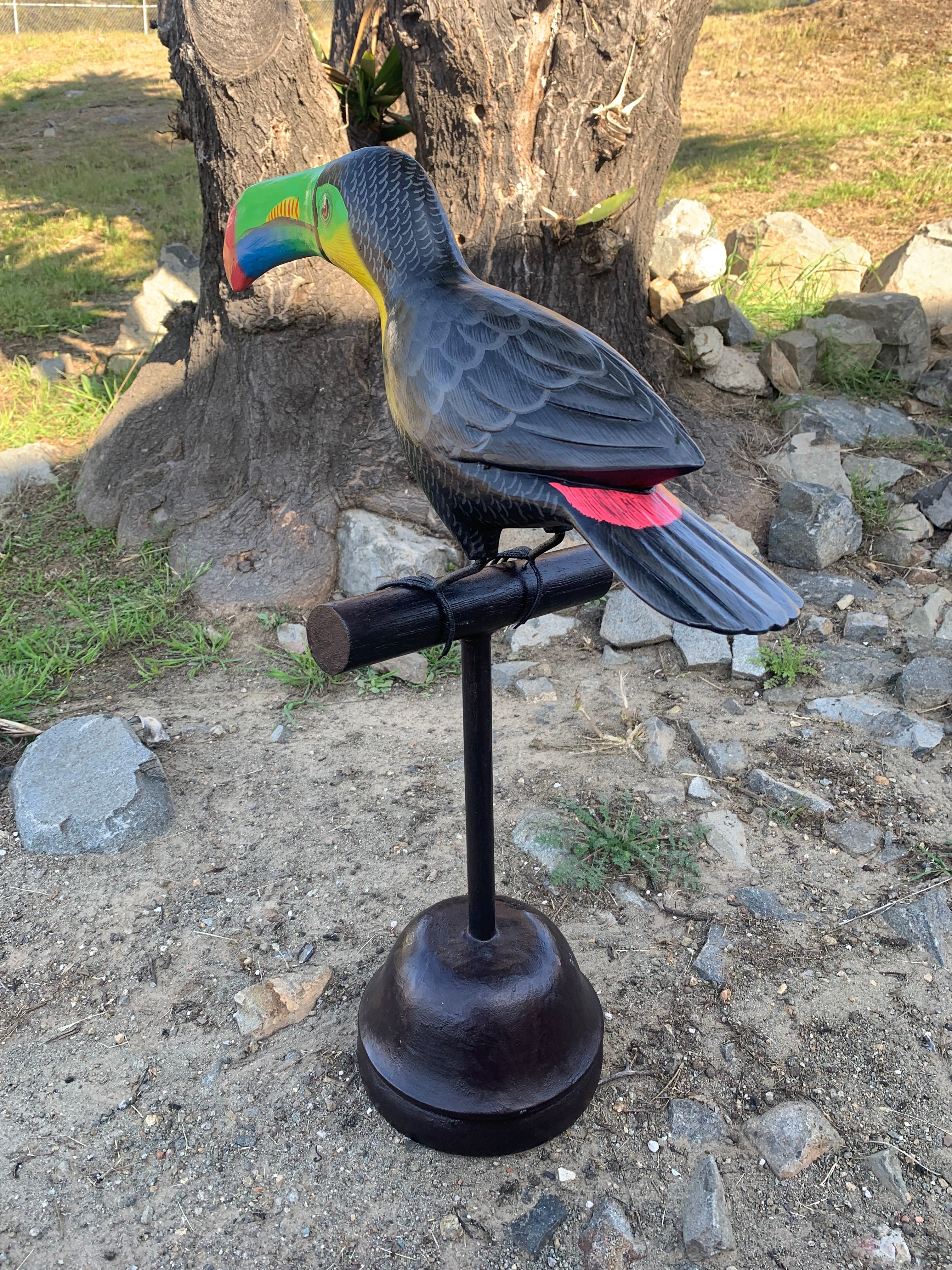 Brightly colored toucan statue perched on a black metal stand in a rocky outdoor yard.