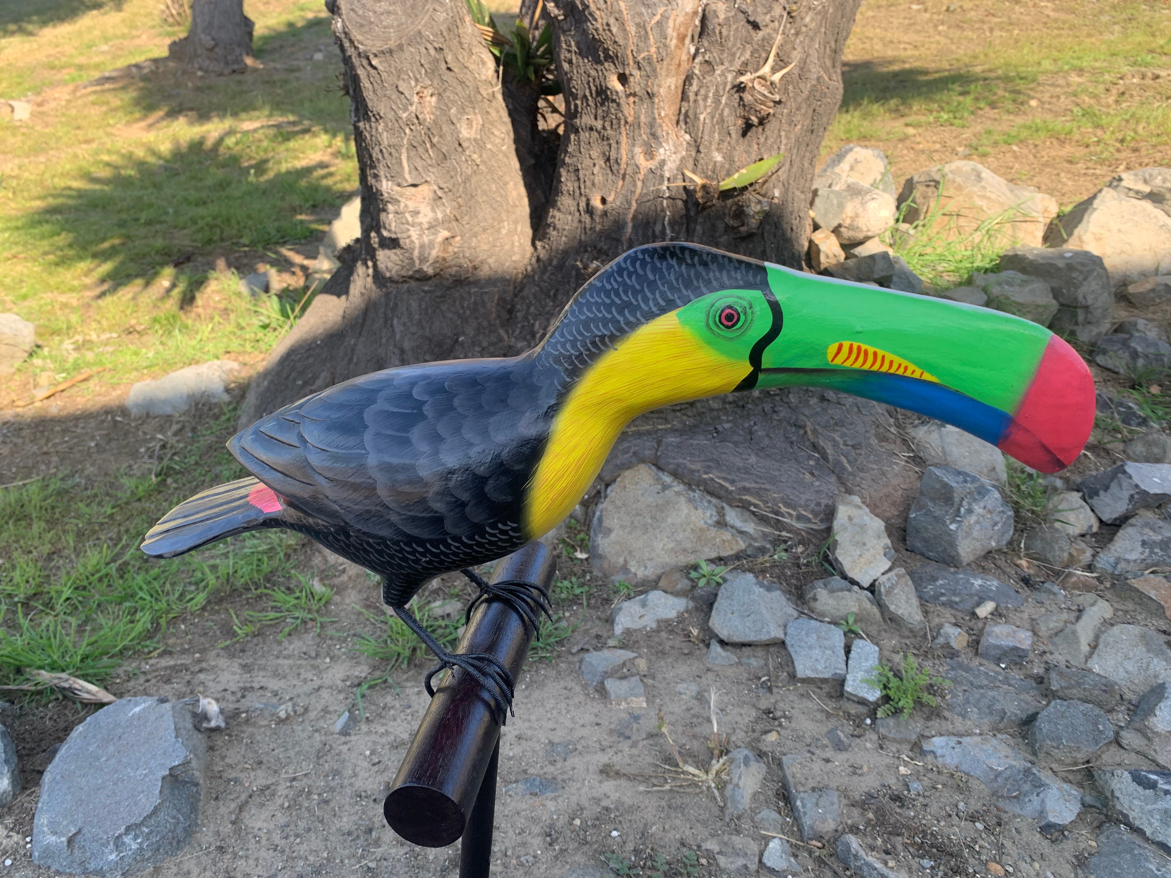 Colorful toucan sculpture perched on a stand outdoors among rocks and a tree
