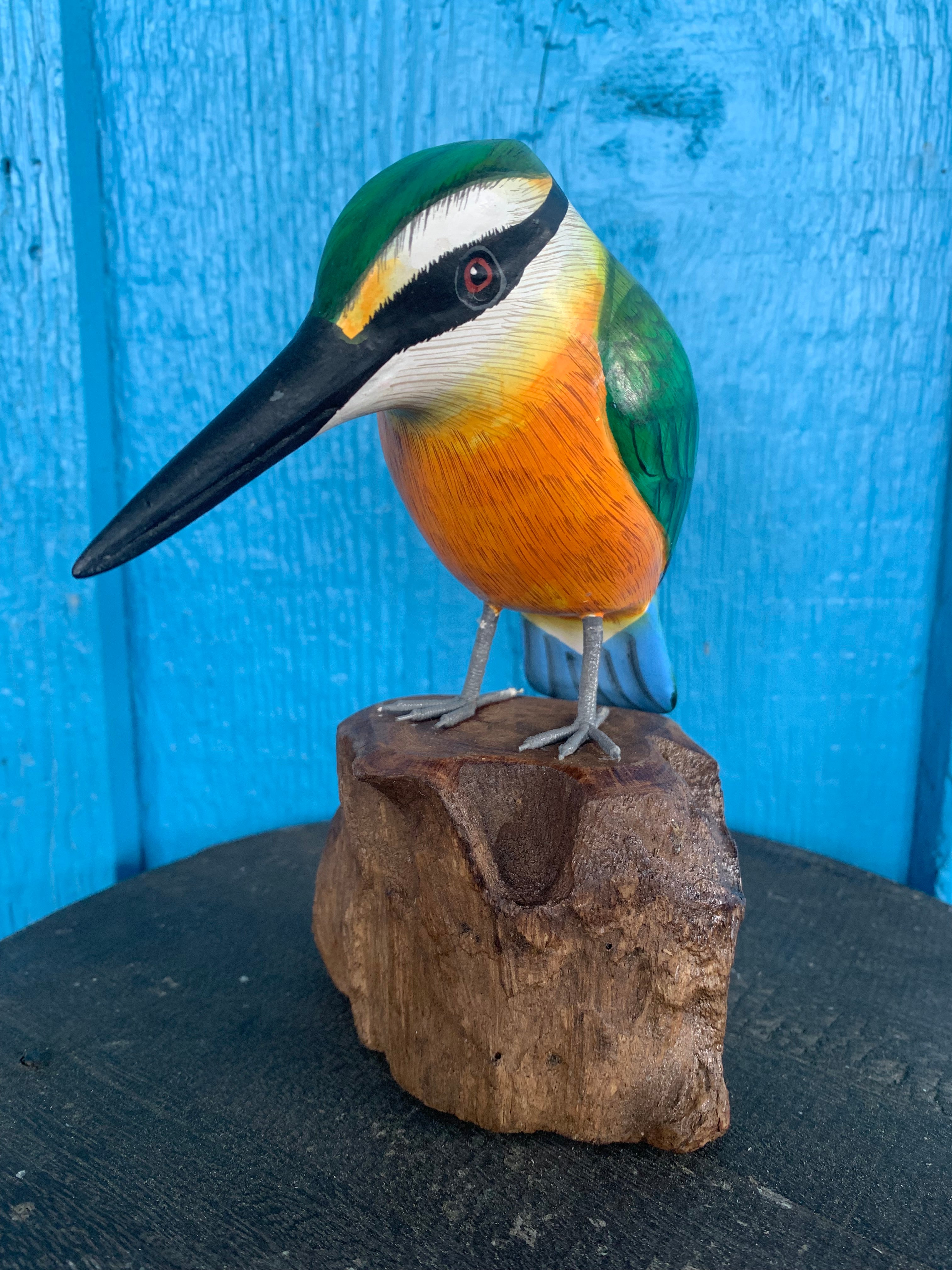 Colorful wooden kingfisher sculpture perched on a log against a blue wall.
