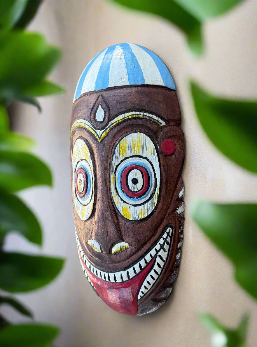 Colorful wooden mask with blue-striped dome, wide circular eyes, and a grinning mouth with tongue showing.