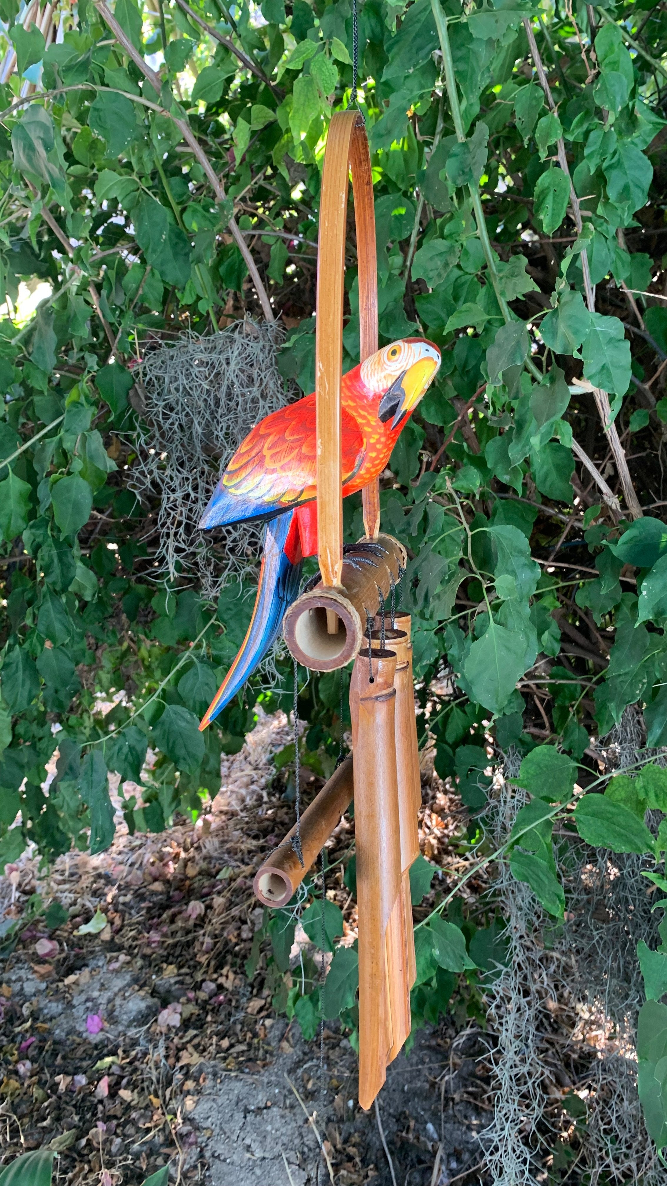 Colorful wooden parrot sculpture hanging on bamboo wind chimes in a lush garden.