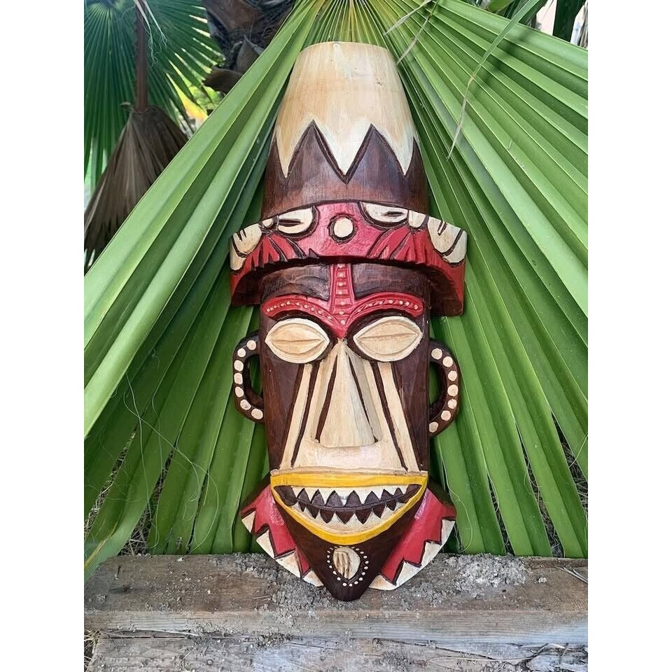Colorful wooden tiki mask carving with red, beige and yellow details among green palm leaves