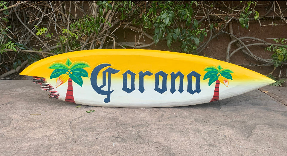 Surfboard with Corona logo and palm trees, yellow gradient top, on pavement.