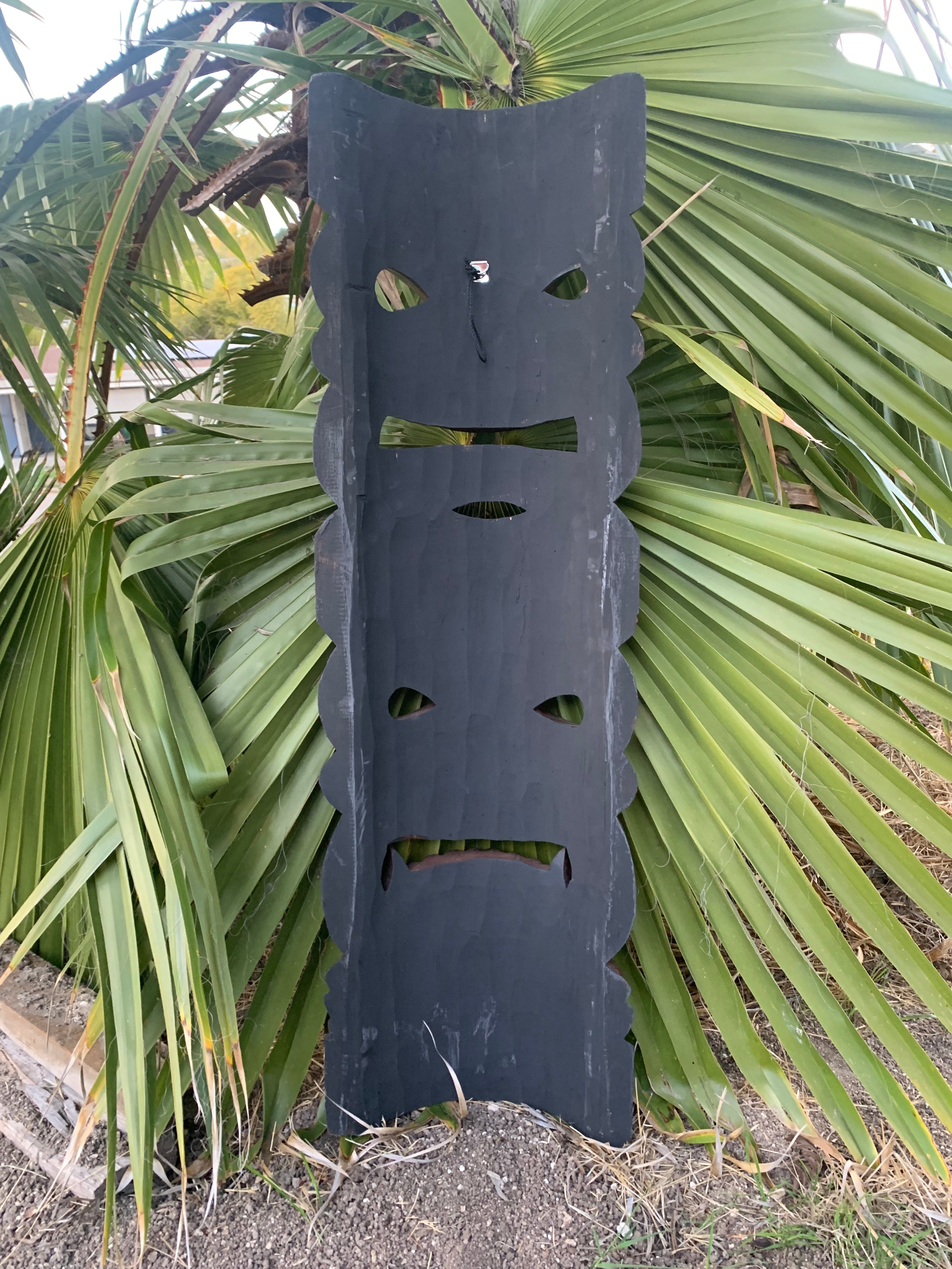 Dark wooden tiki totem with face cutouts leaning against palm fronds
